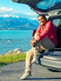 Smiling Man Sits On Open Rear Car Hatch By Lake And Mountain Views